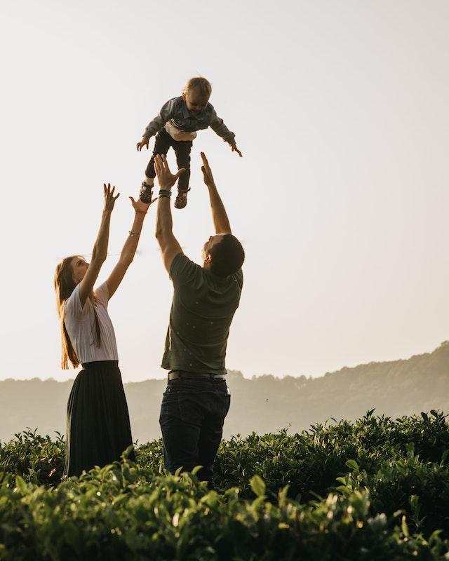 Two adults in a green field, tossing a toddler into the air, arms outstretched to catch
